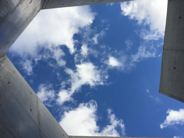 "Pavillon" Blick nach oben durch eine Beton-Öffnung zum blauen Himmel mit weißen Wolkenformationen.