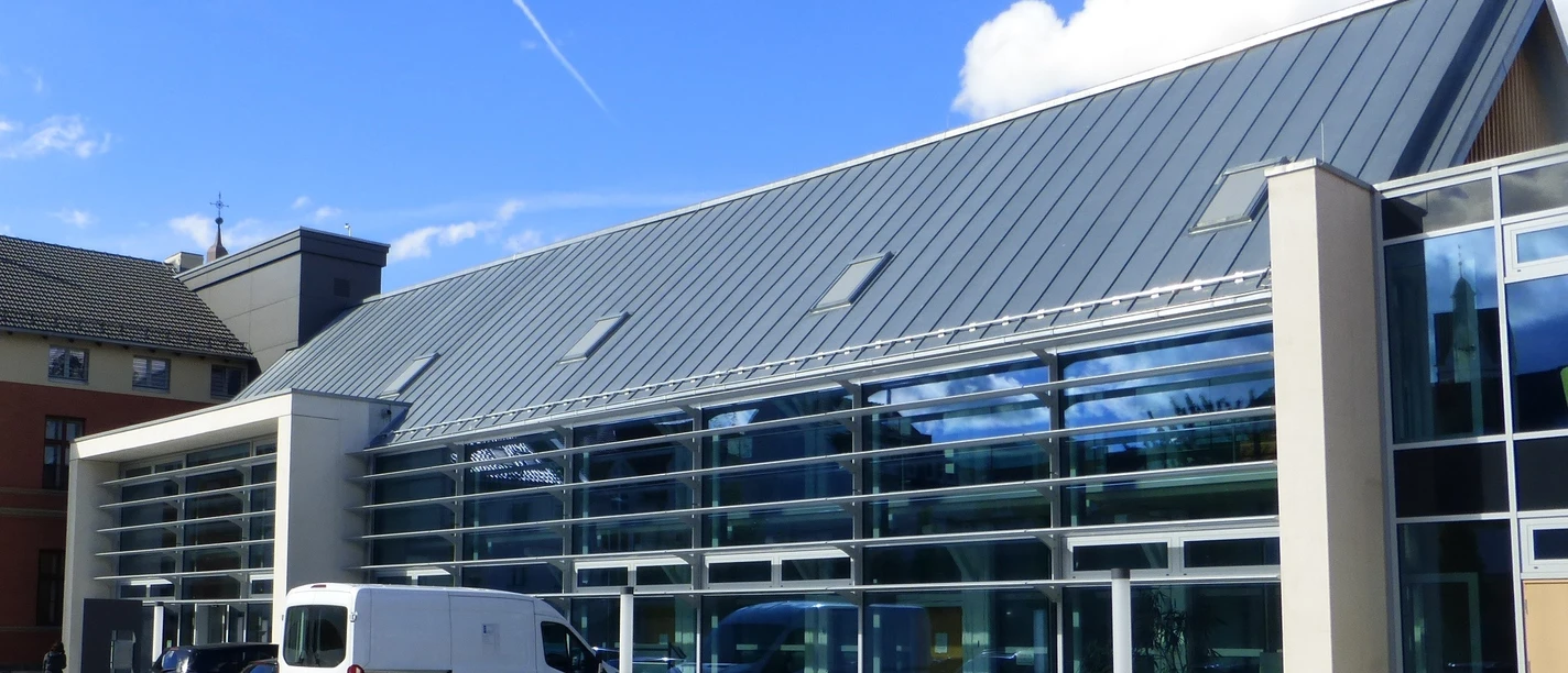 Moderne Glasfassade eines Gebäudes mit geneigtem Metalldach, Autos parken davor, blauer Himmel.