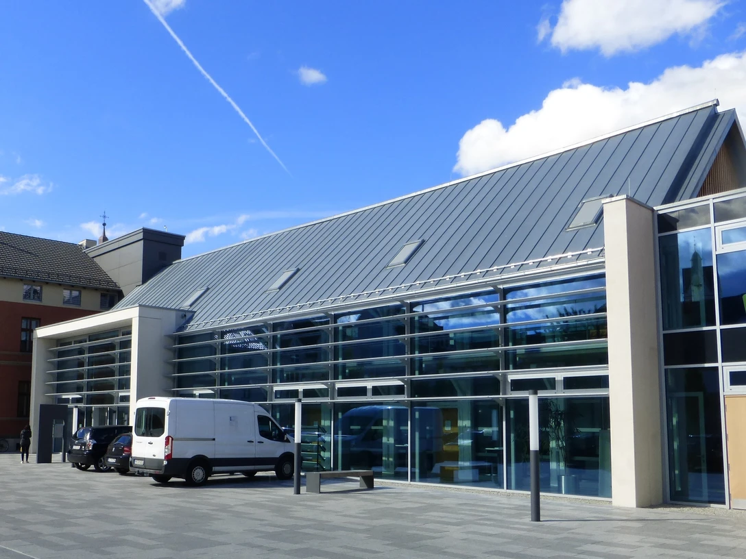 VHS Paderborn Moderne Glasfassade eines Gebäudes mit geneigtem Metalldach, Autos parken davor, blauer Himmel.