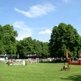 Reitturnier auf dem Schützenplatz Paderborn Reitturnier auf grünem Platz, Hindernisse und Teilnehmer umgeben von üppigen Bäumen, blauer Himmel.