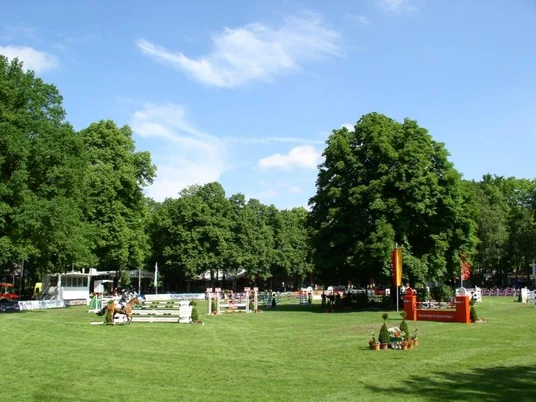Reitturnier auf dem Schützenplatz Paderborn Reitturnier auf grünem Platz, Hindernisse und Teilnehmer umgeben von üppigen Bäumen, blauer Himmel.