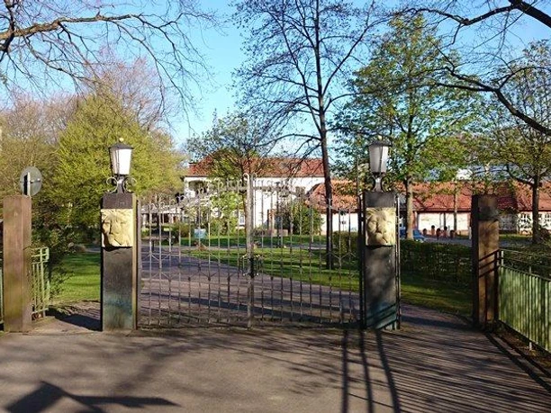 Eingang mit Löwentor zum Schützenplatz Paderborn, umgeben von Bäumen und blauer Himmel im Hintergrund.