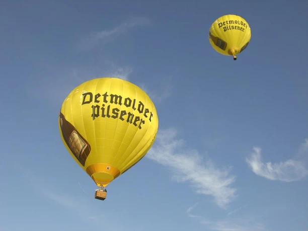 Zwei gelbe Heißluftballons mit der Aufschrift "Detmolder Pilsener" schweben am blauen Himmel.