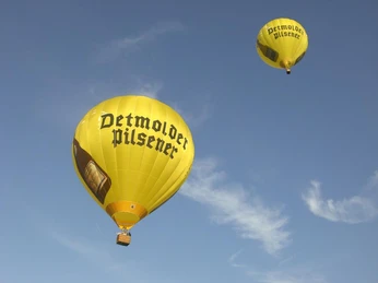 Jede Ballonfahrt ist einzigartig! Zwei gelbe Heißluftballons mit der Aufschrift "Detmolder Pilsener" schweben am blauen Himmel.
