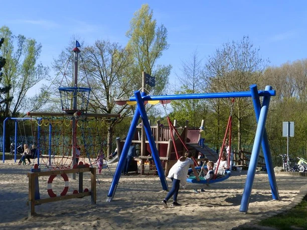 Spielplatz am Padersee mit Klettergerüst, Schaukeln und Sandfläche, umgeben von Bäumen im Hintergrund.