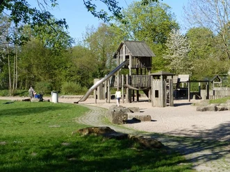 Spielplatz mit Holzklettergerüst und Rutsche, umgeben von grünen Bäumen und Wiese.