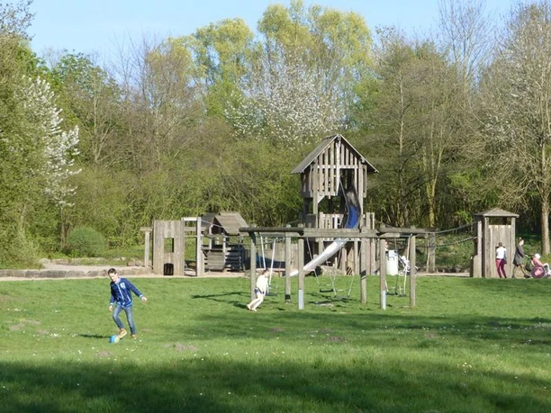 Spielplatz mit Holzspielgeräten, Schaukel und Kletterturm, umgeben von grüner Wiese und Bäumen.