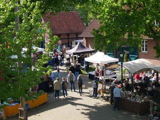 Besucher schlendern durch einen sonnigen Kunsthandwerkermarkt, umgeben von Bäumen und historischem Backstein.