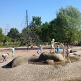 Kinder spielen auf einem Spielplatz mit Wasserspielen, Klettergerüst und Bäumen im Hintergrund.
