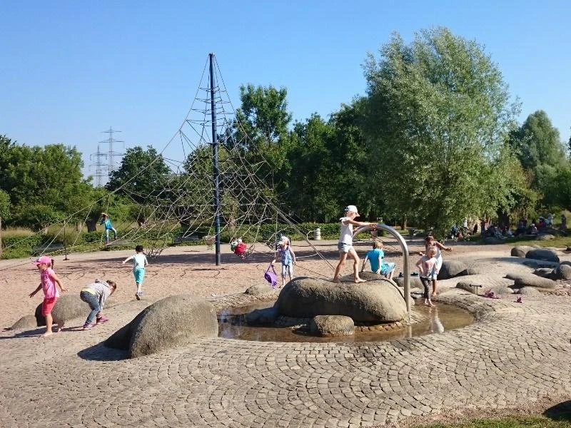 Kinder spielen auf einem Spielplatz mit Wasserspielen, Klettergerüst und Bäumen im Hintergrund.