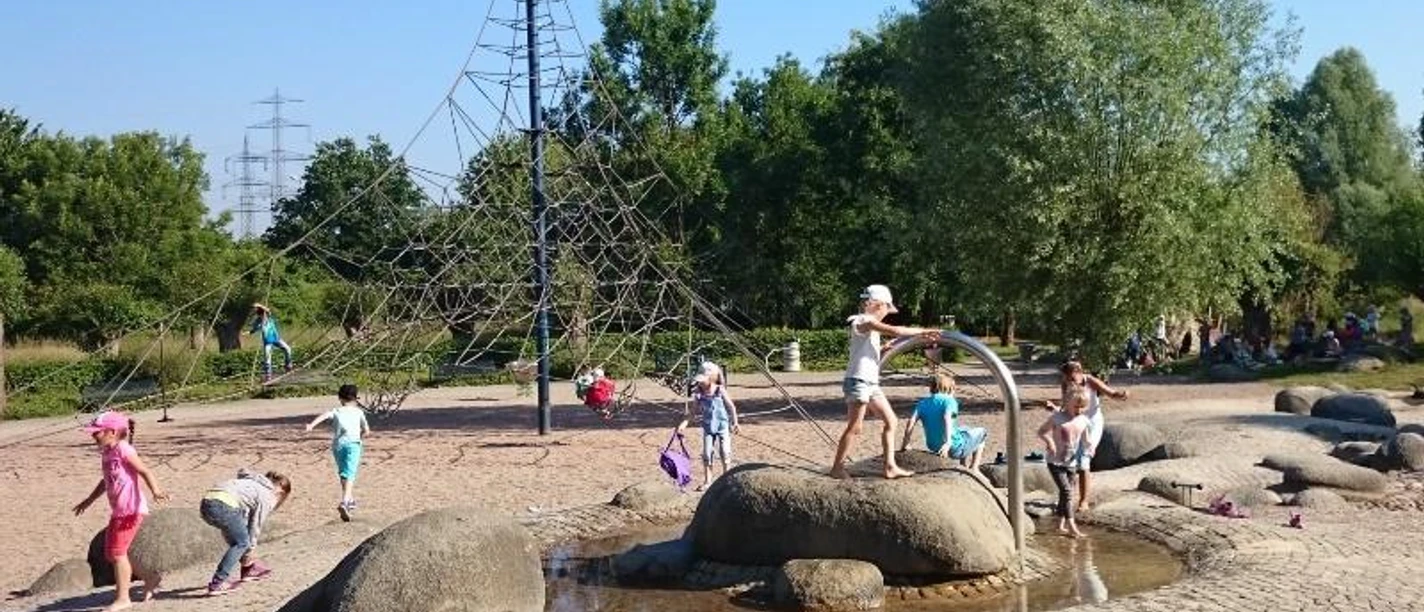 Spielplatz im Schloß- und Auenpark Kinder spielen auf einem Spielplatz mit Wasserspielen, Klettergerüst und Bäumen im Hintergrund.