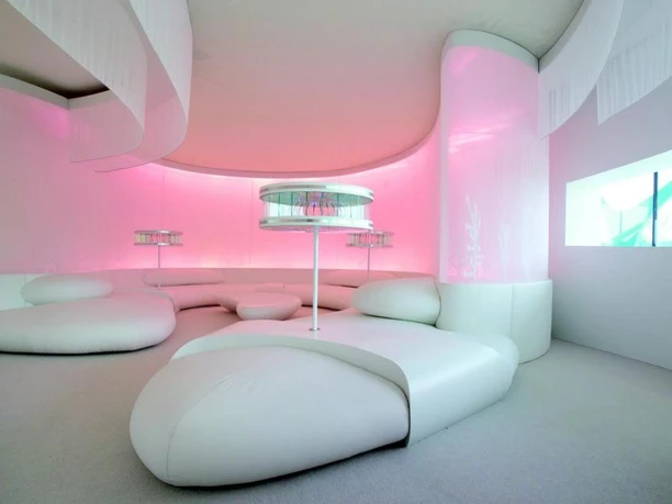 Leonardo glass cube Futuristisch gestalteter Raum mit weißen, geschwungenen Sitzmöbeln und sanfter rosa Beleuchtung.