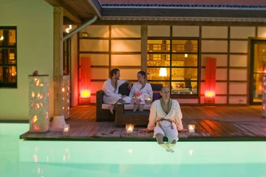 Bali Therme: Atrium Drei Personen in Bademänteln entspannen bei Kerzenlicht auf einer Terrasse neben einem beleuchteten Pool.