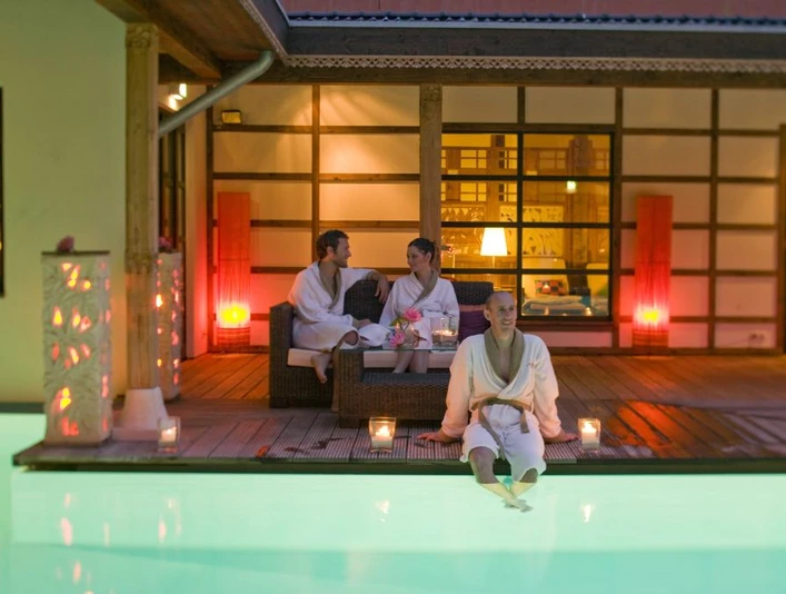 Bali Therme: Atrium Drei Personen in Bademänteln entspannen bei Kerzenlicht auf einer Terrasse neben einem beleuchteten Pool.