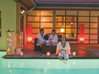 Sauna Atrium in der Bali Therme Personen in Bademänteln entspannen auf einer Terrasse mit dampfendem Wasserbecken im Vordergrund.