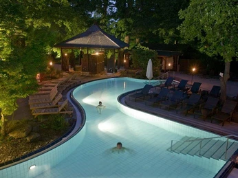 Bali Therme: Saunagarten Bei Dämmerung schwimmen zwei Personen im beleuchteten Außenpool, umgeben von Holzbänken und Bäumen.