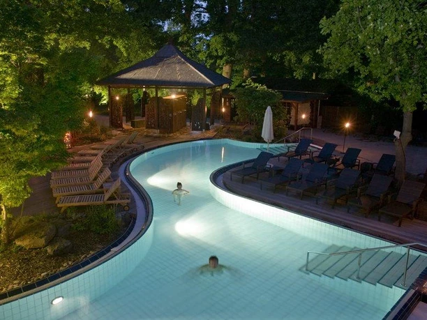 Bali Therme: Saunagarten Bei Dämmerung schwimmen zwei Personen im beleuchteten Außenpool, umgeben von Holzbänken und Bäumen.