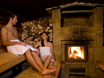 Erdsauna Holzverkleidete Sauna mit Kamin, zwei entspannte Personen, in Handtücher gehüllt, vor gestapeltem Holz.