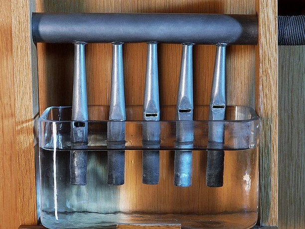Wasserpfeifen Fünf Metallrohre hängen über einem mit Wasser gefüllten Glasbehälter in einem hölzernen Rahmen.