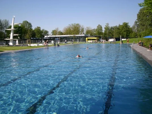 Schwimmer im Freibad schwimmen in einem großen, langen Becken unter blauem Himmel und umgeben von Bäumen.