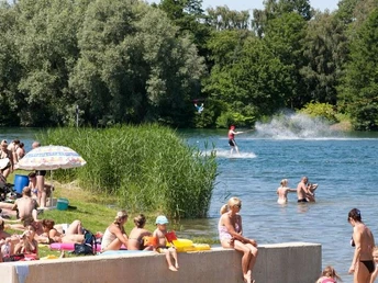 Menschen genießen einen sonnigen Tag am Nesthauser See, während jemand im Wasser Wasserski fährt.