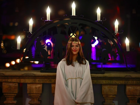 Borchenener Adventsmarkt-Engel Engel in weißem Gewand lächelt vor einem großen, beleuchteten Kerzenständer mit violettem Glanz.