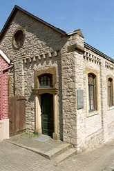 Kleine Synagoge aus hellem Stein mit Spitzgiebel, Rundfenstern und grüner Holztür, lateralansicht.