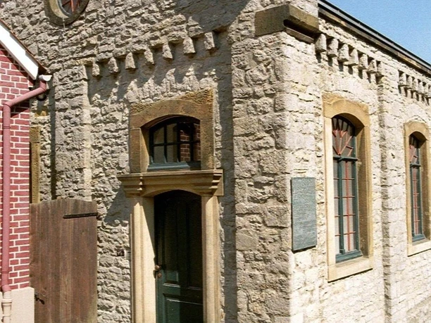 Kleine Synagoge aus hellem Stein mit Spitzgiebel, Rundfenstern und grüner Holztür, lateralansicht.