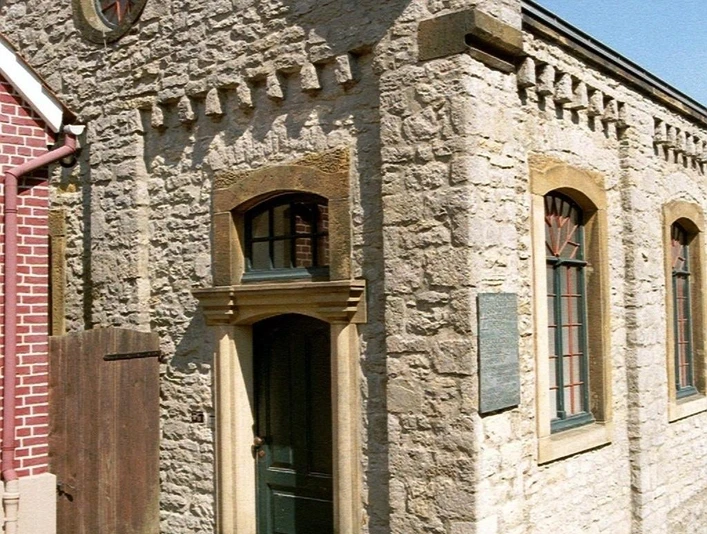 Kleine Synagoge aus hellem Stein mit Spitzgiebel, Rundfenstern und grüner Holztür, lateralansicht.