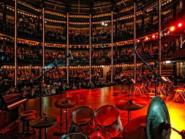 cultura - sparkassentheater an der ems Blick von einer Bühne auf ein vollbesetztes, beleuchtetes Rundtheater mit Orchesterinstrumenten.
