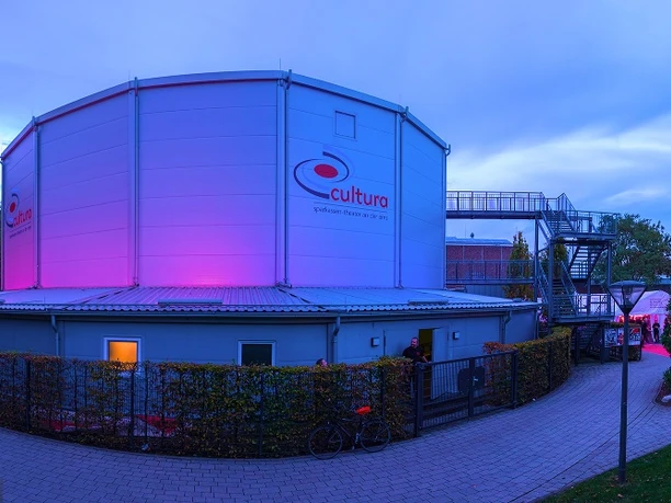 Cultura Rietberg Außenansicht Ein beleuchteter, runder Veranstaltungsbau im Park, umgeben von Bäumen bei Dämmerung.