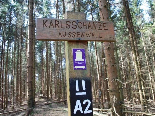 Karlsschanze Holzschild mit der Aufschrift "Karlsschanze Außenwall" im Nadelwald mit Lichtdurchflutung.