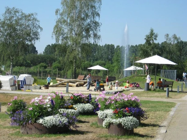 Wasser-Matsch-Spielplatz Spielplatz im Grünen mit Blumenbeeten, Sandkasten und Kindern beim Spielen; weiße Sonnenschirme.