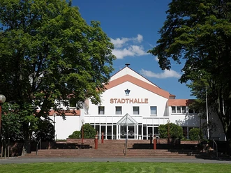 Stadthalle Brakel Große weiße Stadthalle mit rotem Schriftzug, umgeben von Bäumen, unter blauem Himmel.