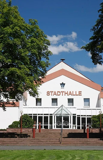 Stadthalle Brakel Große weiße Stadthalle mit rotem Schriftzug, umgeben von Bäumen, unter blauem Himmel.