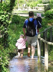 Barfusspark Lienen Ein Mann mit zwei kleinen Kindern läuft barfuß durch einen Wasserweg mit üppigem Grün drumherum.