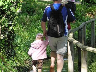 Barfusspark Lienen Ein Mann mit zwei kleinen Kindern läuft barfuß durch einen Wasserweg mit üppigem Grün drumherum.