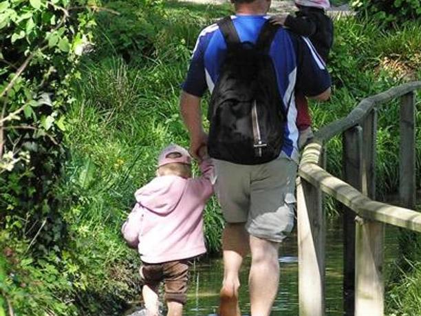 Barfusspark Lienen Ein Mann mit zwei kleinen Kindern läuft barfuß durch einen Wasserweg mit üppigem Grün drumherum.
