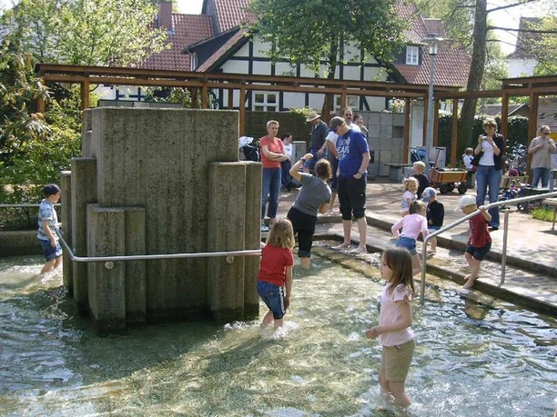 Lienen Barfusspfad Kinder und Erwachsene spazieren über einen Wasserparcours, umgeben von Grünanlagen und Fachwerkhäusern.