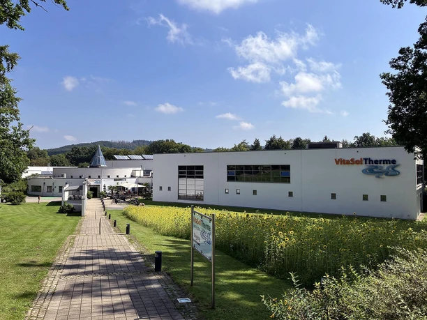 Außenansicht der VitaSol Therme mit Weg, Wiese und Sonnenblumen unter blauem Himmel