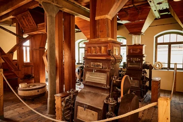 Historische molen met oude houten en metalen machines, omringd door houten balken en touwen, in een ruimte met warm licht en ramen op de achtergrond.