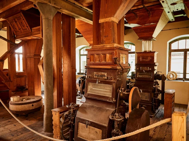 Historic mill with old wooden and metal machines, surrounded by wooden beams and ropes, in a room with warm light and windows in the background.