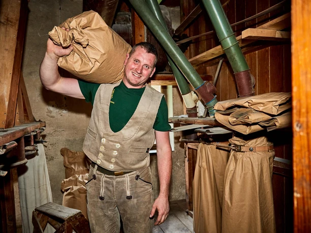 A man in traditional work clothes carries a large brown sack on his shoulder in a rustic mill with pipes and sacks.
