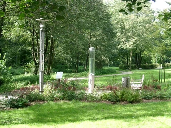 Grüne Parklandschaft mit zentraler moderner Wassersäule umgeben von Bänken und Bäumen.