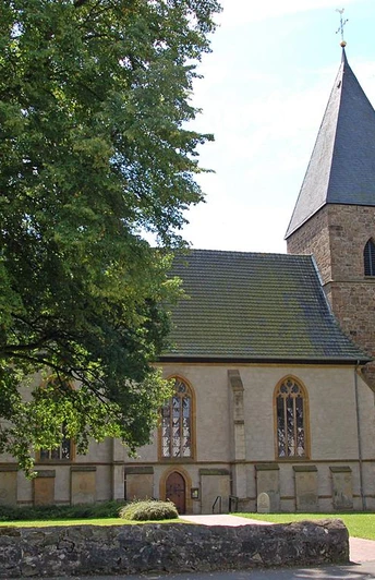 Stiftskirche in Stift Quernheim mit steinernem Glockenturm und umgebenden Laubbäumen im Sommer.
