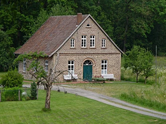 Ein traditionelles Backsteinhaus vor einem grünen Wald, mit gepflegtem Rasen und zwei weißen Bänken.