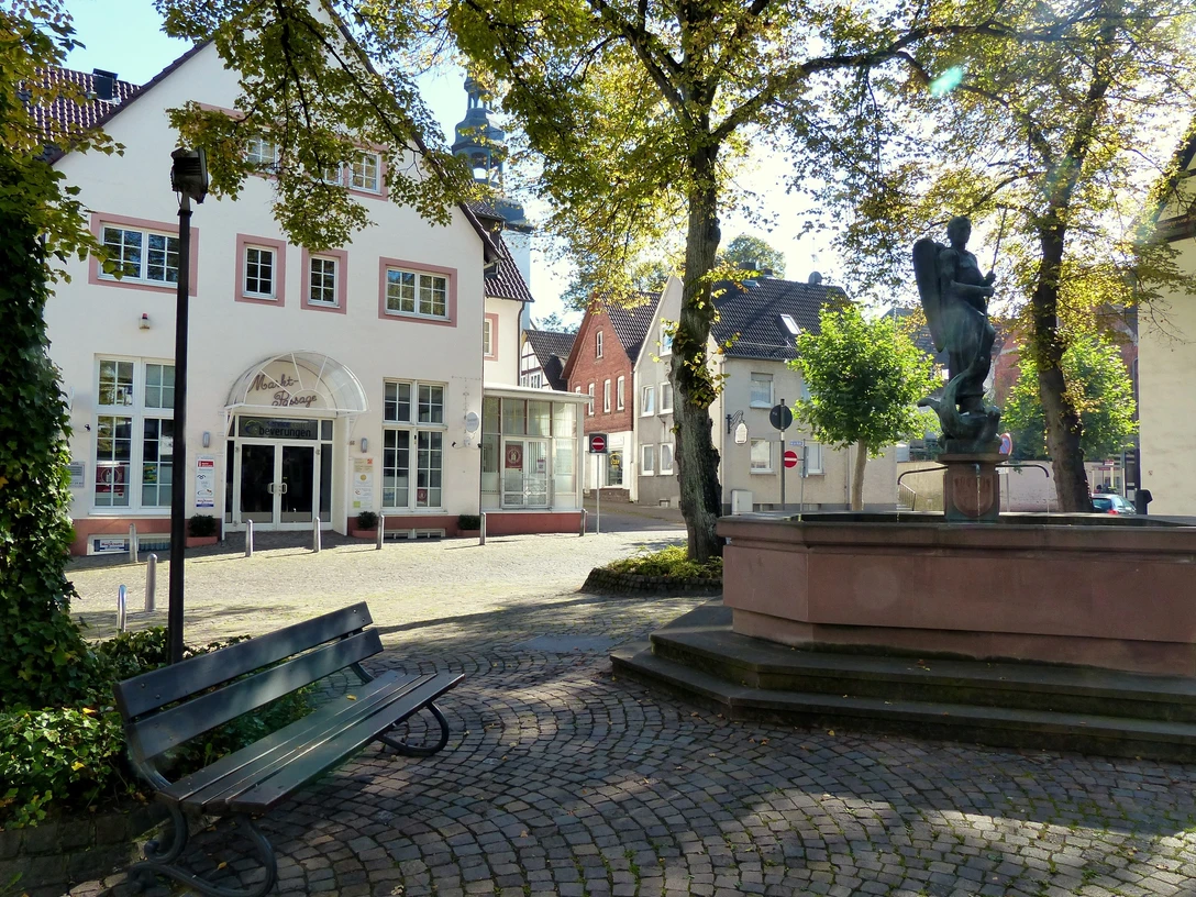 Ein historisches Haus mit Fachwerk an einem gepflasterten Platz, umgeben von Bäumen und einem Brunnen.