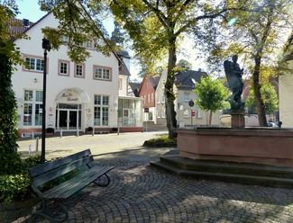 Service-Center + Brunnen Ein historisches Haus mit Fachwerk an einem gepflasterten Platz, umgeben von Bäumen und einem Brunnen.