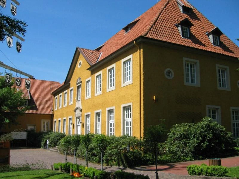 Hilter - Rathaus Gelbes Rathausgebäude mit roten Ziegeldächern, umgeben von gepflegten Sträuchern und Bäumen.