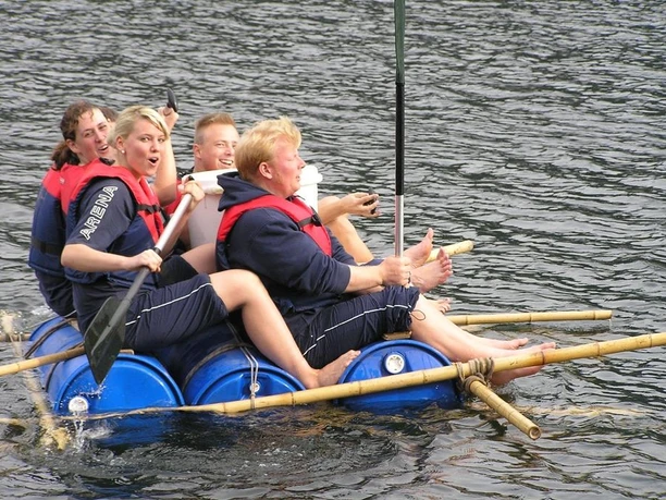 Eine Gruppe junger Menschen paddelt lachend auf einem selbstgebauten Floß über einen See.